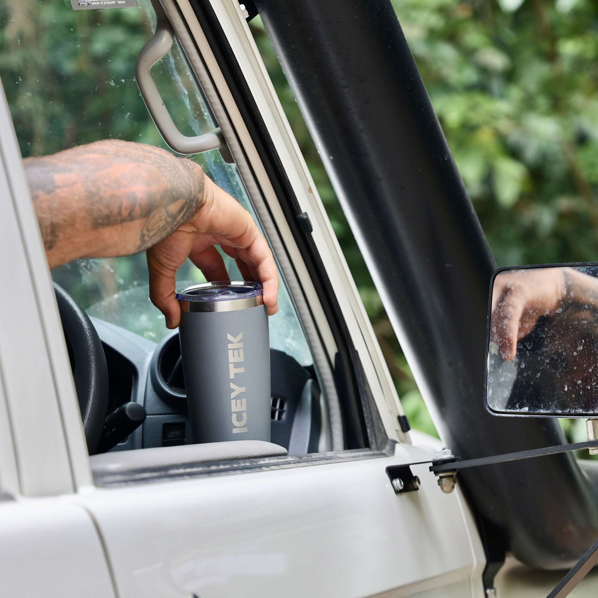 Person holding a cup near a car window with a side mirror reflecting their hand.