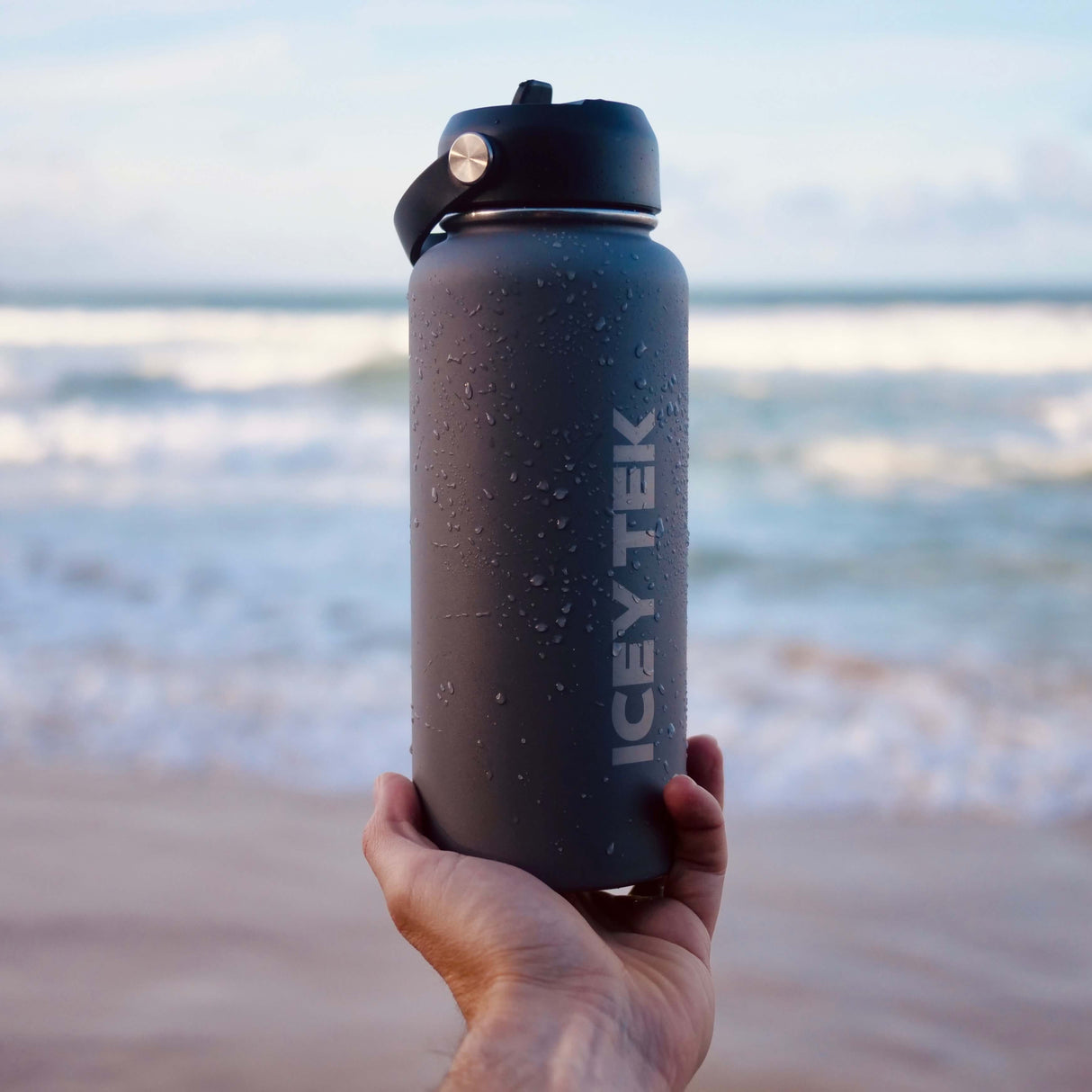 Hand holding an ICY TEK water bottle with a beach background