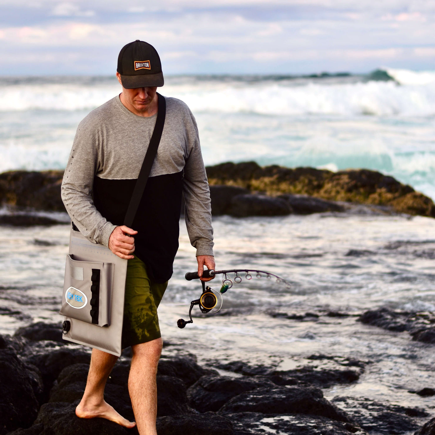 Man standing on rocks by the ocean holding a fishing rod and tackle box.