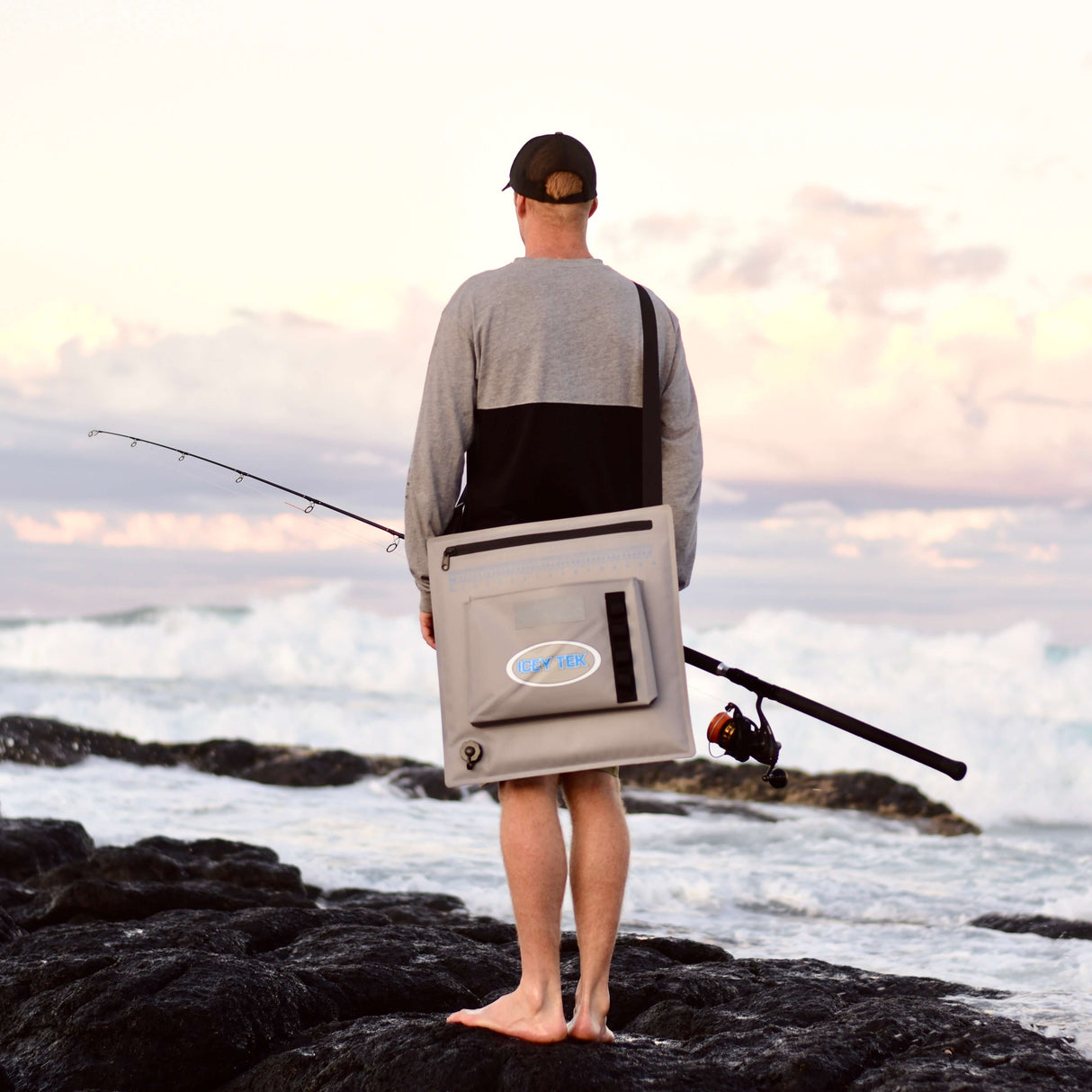 Person standing on rocks by the ocean with a cooler and fishing rod, wearing a cap and gray jacket.