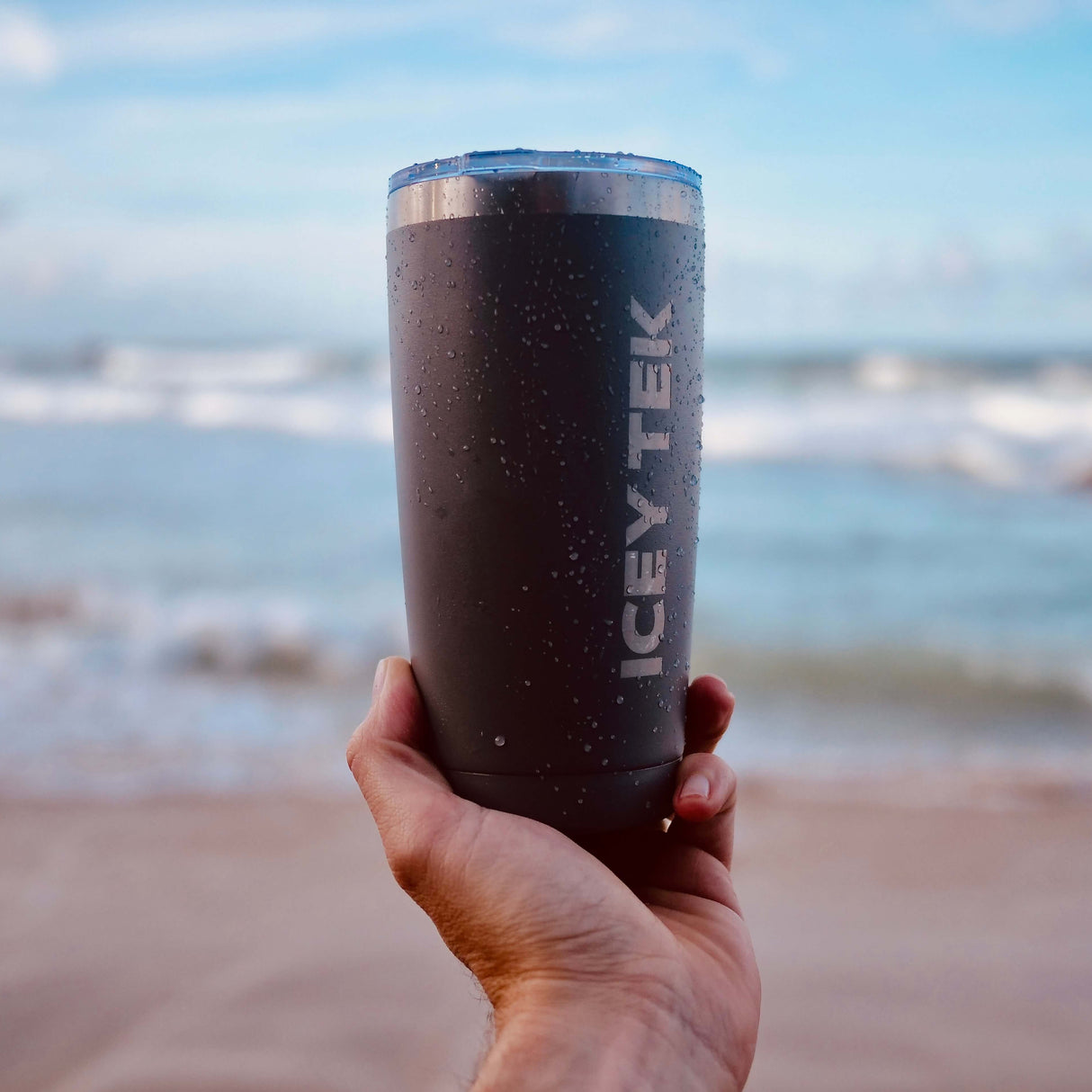 Person holding an 'Icey Tek' tumbler by the beach